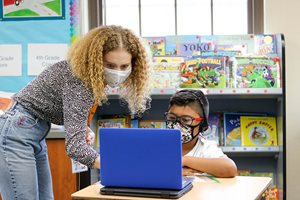 A masked adult assisting a child at a laptop